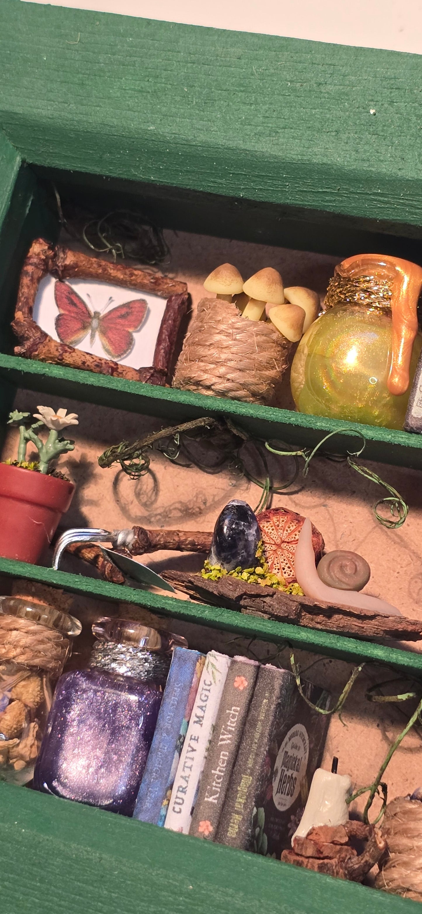 A close-up of a green shadow box containing whimsical fairy tale and nature-themed decorations, including a glass bottle, small books, a butterfly print, and dried botanicals.