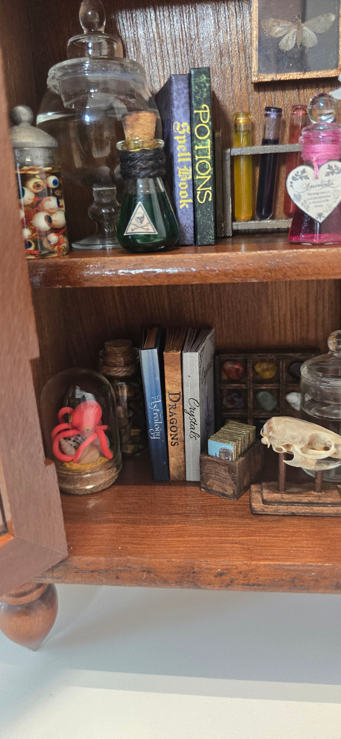 A close-up of a wooden shelf or cabinet containing themed spellbook-style books, glass bottles, beads, and small decorative objects, including a book titled 'Potions Spell Book'.